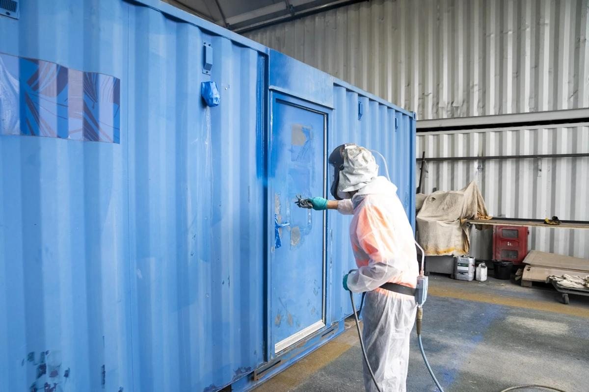 Detail of industrial worker using spray gun to apply powder coating on metal structure inside factory booth.